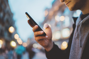 Side view image of young hipster man using modern smartphone outdoors, man typing an sms message while walking at evening city streets, social networks concept