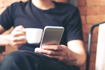 A man using smart phone and holding coffee cup in modern cafe with brick background