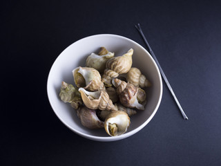 Whelks (sea snails) in white bowl with snail fork isolated on black background