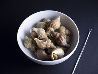 Whelks (sea snails) in white bowl with snail fork isolated on black background