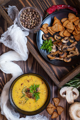 Pea soup with mushrooms in black plate. Fried mushrooms. Dried pea, pepper in glass jars.  Fresh parsley and rosemary. Rye crackers. Dark wooden table. Toned.