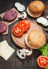 Homemade hamburger ingredients. Raw minced beef, fresh bun, slice of cheese, tomato, onion rings, lettuce on wood background