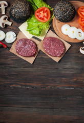 Homemade hamburger ingredients. Raw minced beef, fresh black bun, slice of cheese, tomato, onion rings, lettuce on wood background