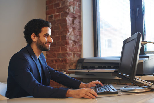 An Arab man in a jacket sitting in the workplace at the computer and typing on keyboard in office