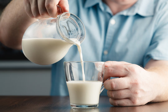 Close Up Of Man Hand Pouring Milk From Jug To Glass On A Rustic Wooden Table