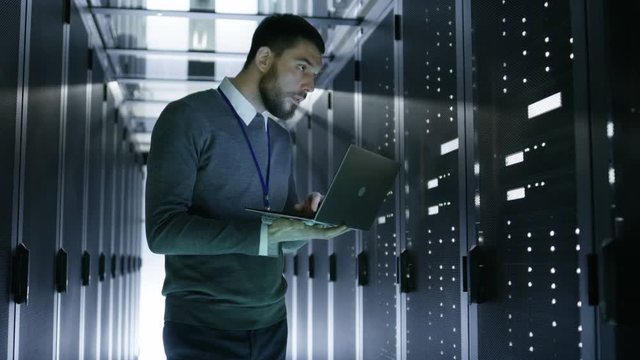 IT Technician Walks and Works on a Laptop in Big Data Center full of Rack Servers. He Runs Diagnostics and Maintenance Stets up System. Shot on RED EPIC-W 8K Helium Cinema Camera.
