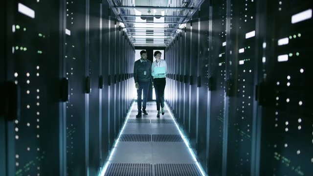 Female IT Technician and Male Server Engineer Walk and Discuss Settings of a Working Data Center. Woman Holds Laptop.  Shot on RED EPIC-W 8K Helium Cinema Camera.