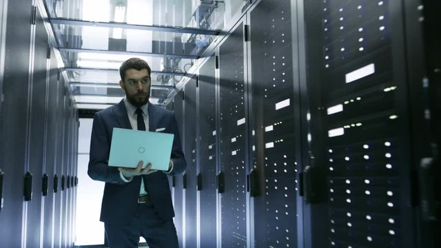 IT Engineer Walking in Data Center full of Rack Servers, He Holds Laptop and Runs Diagnostics. Shot on RED EPIC-W 8K Helium Cinema Camera.