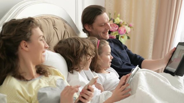 Closeup Portrait of Young Family Lying on the Bed in the Morning and Using Their Gadgets. Parents and Kids Wearing Pajamas. Handsome Father is Showing Something on His Tablet, Everyone is Laughing.