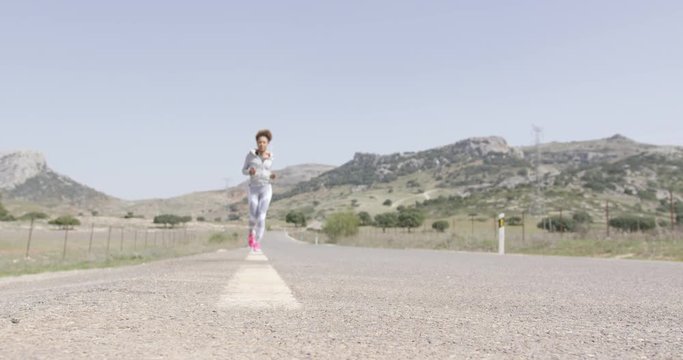 Young fit female wearing sportive clothing running down road among view of beautiful mountains in summer time.