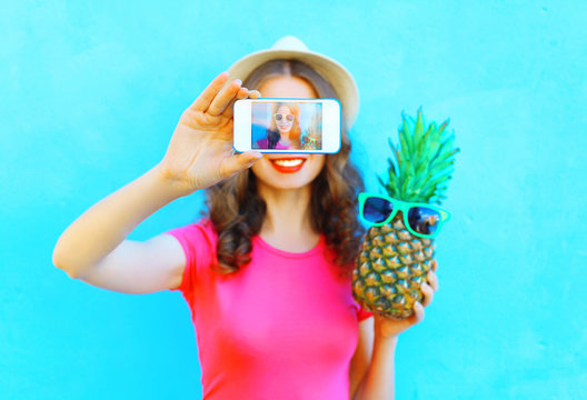 Fashion woman with pineapple taking picture self portrait on smartphone over colorful blue background screen closeup
