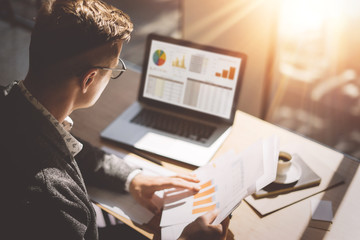 Young finance market analyst in eyeglasses working at sunny office on laptop while sitting at wooden table.Businessman analyze document in his hands.Graphs and diagramm on notebook screen.Blurred. - 141426532
