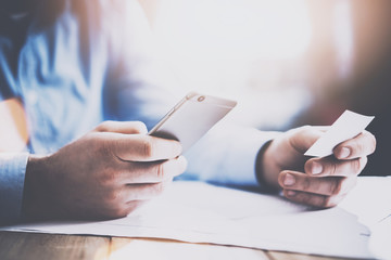 Closeup view of businessman holding business card and smartphone on hands while sitting at the wooden table.Horizontal, blurred background.