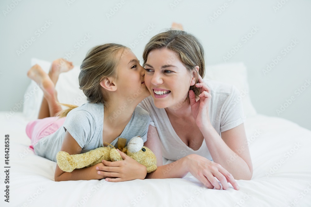 Wall mural Adorable girl kissing her mother lying on a bed