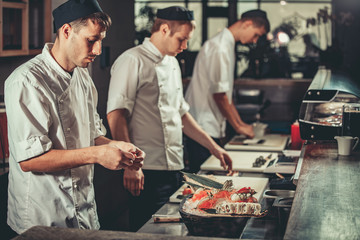 Three young white chefs dressed in white uniform decorate ready dish in restaurant. They are...