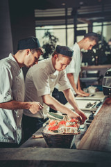 Three young white chefs dressed in white uniform decorate ready dish in restaurant. They are working on maki rolls. Preparing traditional japanese sushi set in interior of modern professional kitchen