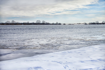 The winter river, covered with snow and ice by the shore, under the blue sky and trees on the horizon