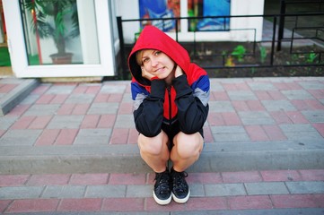 Portrait of a young smiling girl in a sports jacket with a hood, sitting on the steps of porch of a small shop, elbows on bare knees