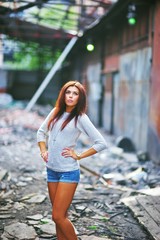 Young beautiful girl the brown-haired woman in a jumper and denim shorts posing at abandoned workshops in summer day.