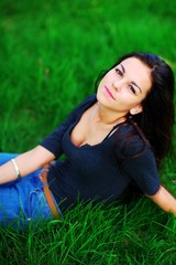 Portrait of young pretty woman brunette with beautiful brown eyes in a dark blouse with a deep cleavage, relaxed on green grass in a Park, closeup.