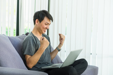 young man sitting on sofa with happy feeling arm up posture when watching laptop computer screen,success celebration concept