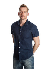 Handsome blonde man in blue shirt and jeans standing against a white background smiling.