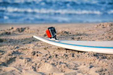 Surfboard with attached action camera lies on the sand on the beach. Action camera is in waterproof case for swimming. Ocean on background