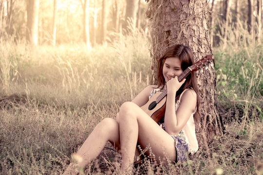 Young asian woman put on earphones listening music and holding acoustic guitalele in the park in vintage color tone