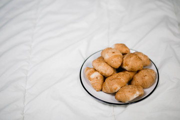 Golden yellow brown yellow tasty delicious sweet croissants, white plate with black stripe in a white bed in the front right foreground with blankets white background