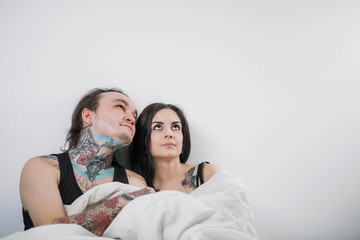 beautiful couple in tattoo in black shirts sitting on the couch looking up under a blanket in copyspace on white background