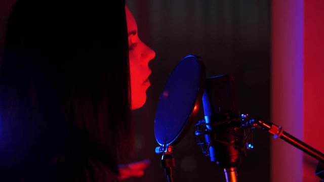 A young woman sings a song in a microphone in a recording studio under Neon light and a smoke machine