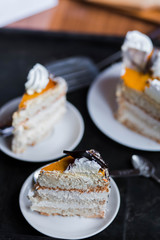 a beautiful delicious cake and the pieces in three white saucers beige yellow on cutting Board on dark black background close-up
