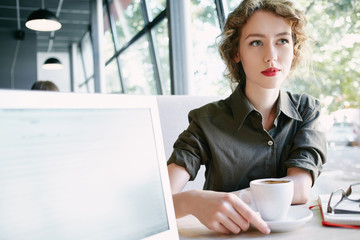 joyful smiling woman sitting in a cafe near the open laptop at a table made of wood . in the background a bright window with bright daylight