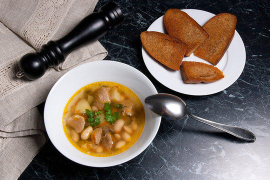 Bean soup in white plate with metal spoon, several toast on white plate on a black stone background.
