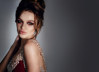 Beautiful, sexy brunette girl with big lips, hair and makeup in a long burgundy dress with precious stones. In a staged on a gray background, a place for advertising