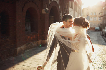 Cheerful wedding couple kisses under the veil in halo of evening light