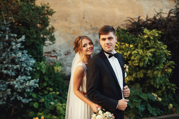 Pretty blonde bride hugs handsome groom from behind standing before the bushes