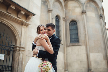 Groom holds tender bride in his arms posing before old white church