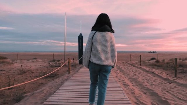 Back view on casual dressed brunette girl in warm sweater and jeans walks on wooden boardwalk to the sand beach at beautiful sunset with pink cyan sky and sits at the end.