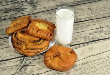 Breakfast. Glass with milk, plate with baking. One bun lies separately. Wooden background