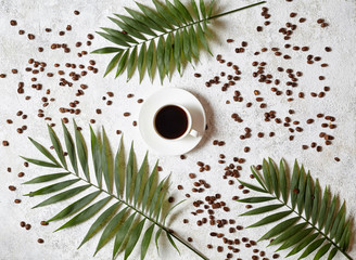 Cup of black espresso on a white concrete background with scattered coffee beans and palm branches. Rest in warm tropical countries concept. Flat lay drink composition.