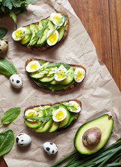 Sandwich, toast with avocado, spinach, guacamole and quail eggs on parchment and dark wooden table. Healthy breakfast lunch concept. Flat lay food composition.