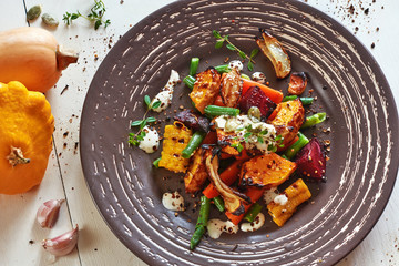 Warm salad with roasted vegetables (pumpkin, carrots, beetroot, green bean, onion) with spices and cream sauce on a white wooden table. 