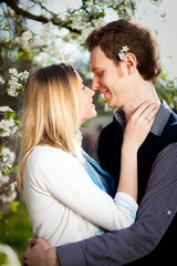 Tender couple in love under blooming cherry tree with white flowers in the park in spring. Happy girl and guy smile and hug outdoor, they are wearing in blue shirt, white jacket.