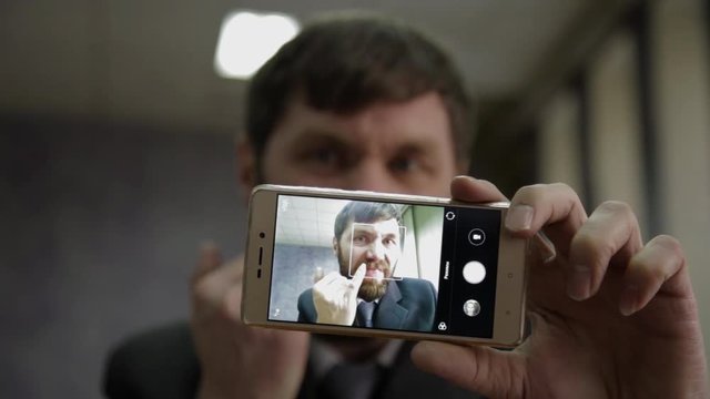 Busy office worker cranks and looking into phone camera. funy businessman picks finger in nose, strokes his beard