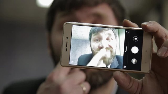 Busy office worker cranks and looking into phone camera. funy businessman picks finger in nose, strokes his beard