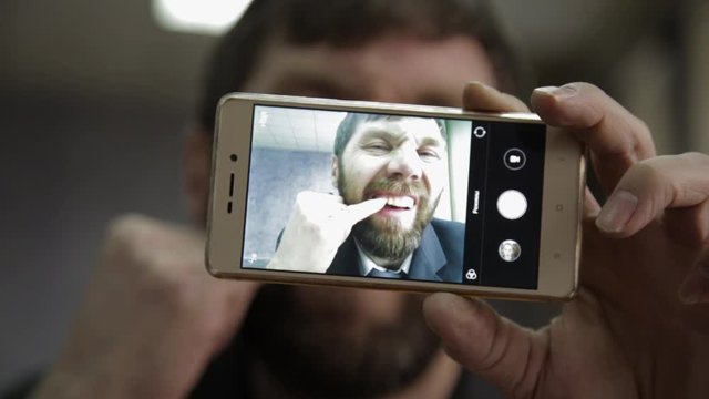 Busy office worker cranks and looking into phone camera. funy businessman picks finger in nose, strokes his beard