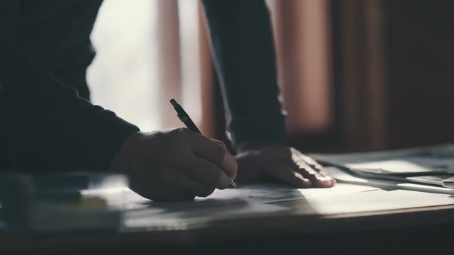 Pensive young handsome man writing in his notebook while sitting at his working place. Close-up: the man bends over the desk and makes notes with a pen, leaning on the table.
