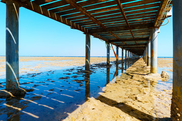 Fototapeta na wymiar View from under the old pier to the shallow sea at low tide. A bright sunny day. Pure blue sky. Clear water. Worn out engineering structure.