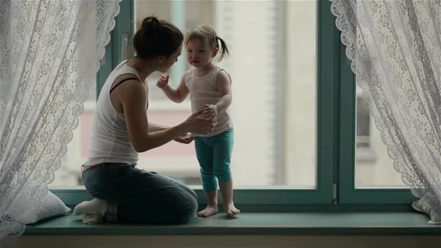 Side View of Loving Mother with Charming Smile and Cute Daughter with Two Ponytails Hugging Each Other next to the Window. Little Girl is Standing on the Windowsill, Amazing Woman is Embracing Her.